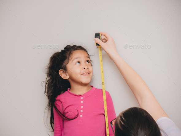 Mother and diverse daughter at home using tape measure to measure height and write it on wall