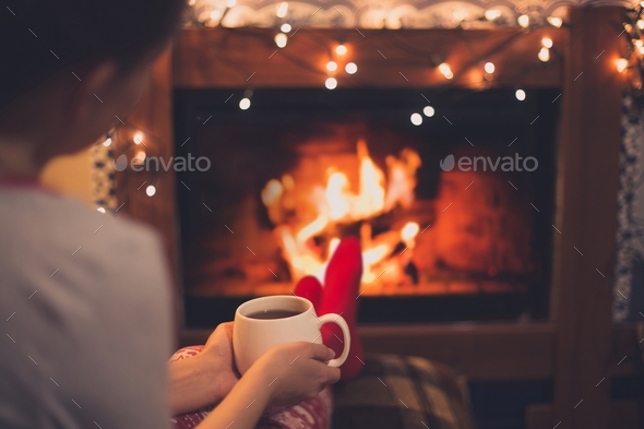 Woman drinking hot tea sitting by fireplace in a cozy house.