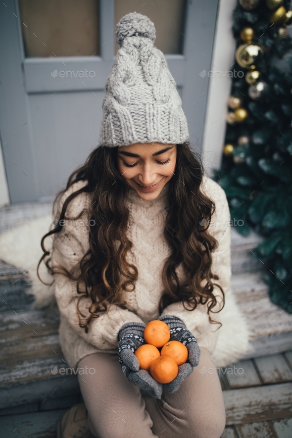 Young woman wearing knitted hat, mittens and sweater sitting on decorated porch. Christmas time