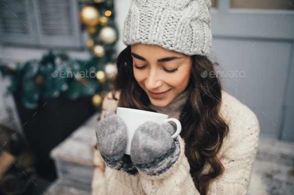 Young woman wearing knitted hat, mittens and sweater drinking tea on decorated porch. Christmas time