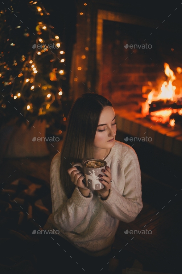 Young woman drinking cocoa with marshmallows sitting by fireplace near Christmas tree in cozy room.