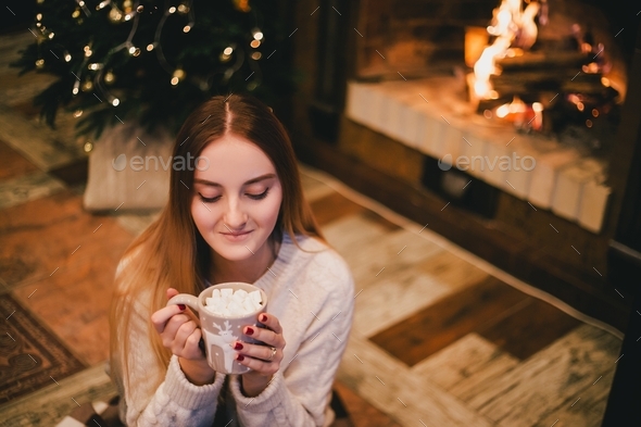 Young woman drinking cocoa with marshmallows sitting by fireplace near Christmas tree in cozy room.