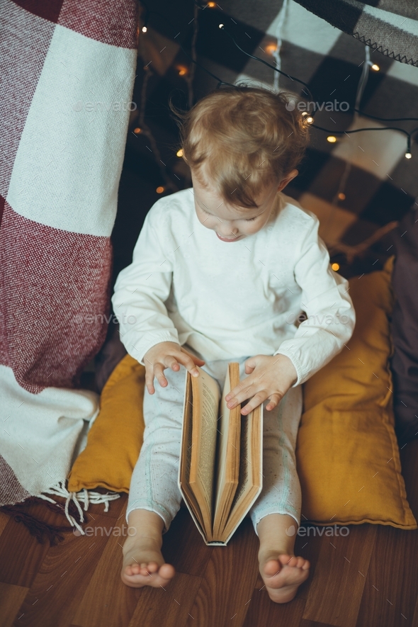 Cute baby reading book in fort made of blankets and garland.