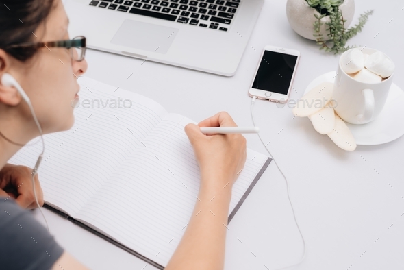 Young millennial woman sitting at a white desk typing on computer while listing to music, school