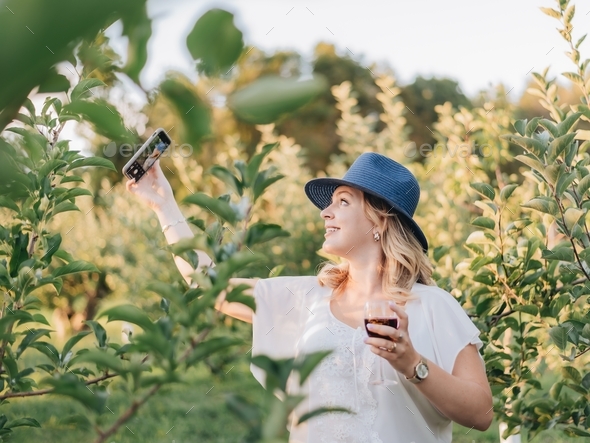 Young millennial girl at an Apple orchard drinking wine and taking a selfie with a smartphone