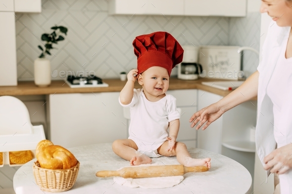 A small beautiful child dressed in a cooking cap sits on the table next to the dough and rolling pin