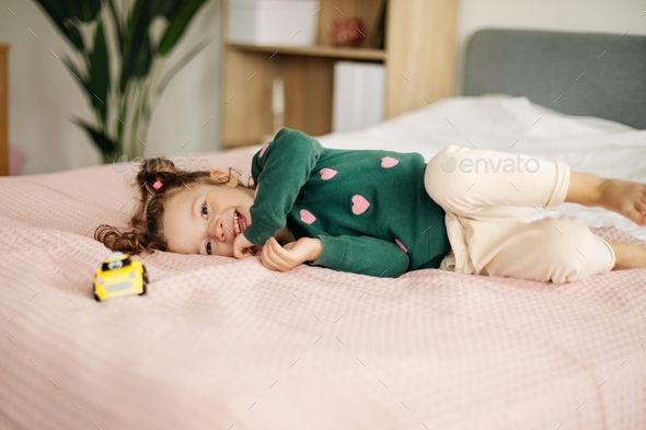 A charming little girl is playing with a car on the bed. Home games, child development. - Stock Photo - Images