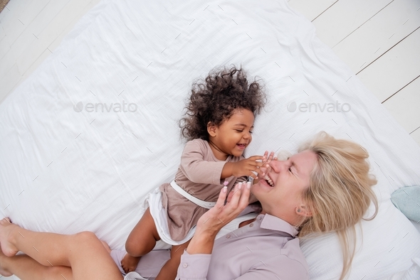 Young Caucasian blond mother tickles little African American daughter. Lie on white bed, having fun