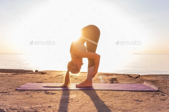 Young blonde woman in sportswear doing yoga asanas Uttanasana pose on the seashore at sunrise