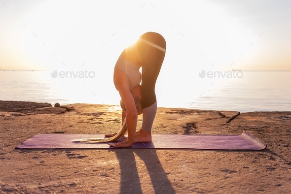 Young blonde woman in sportswear doing yoga asanas Uttanasana pose on the seashore at sunrise