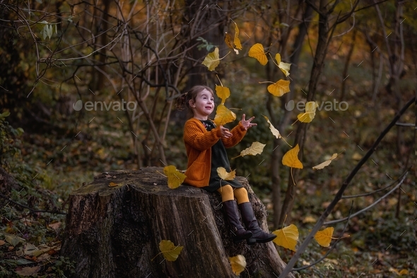 Girl in an orange knitted cardigan, brown boots sits on a stump, throws up yellow poplar leaves