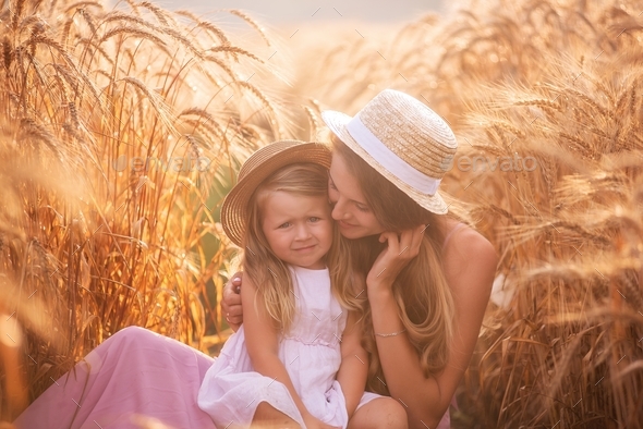 Close-up portrait of mother with daughter in straw hats in wheat field with dew drops.