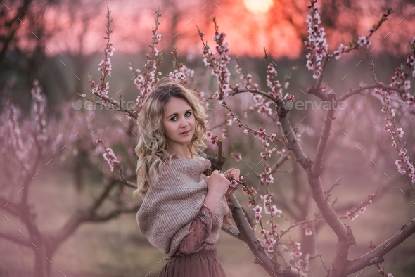 Young beautiful curly blonde woman in brown pleated skirt stands in blooming peach sunset gardens