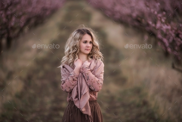 Young beautiful curly-haired blonde woman stands in blooming peach gardens.