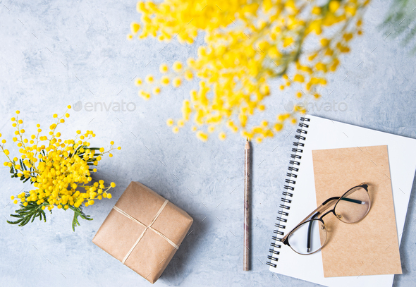 Flat lay with yellow mimosa flower, craft present box, a glasses, a craft notebook on a blue table.