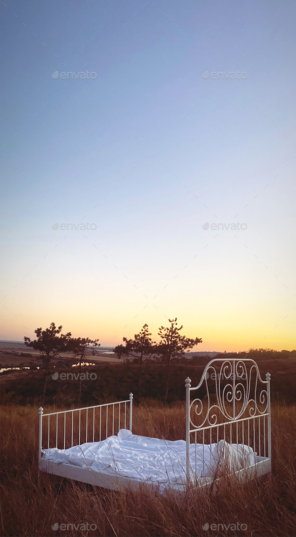 Landscape photo shoot in nature, wrought iron bed with white linen standing in summer evening field