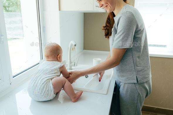 young mom with a baby toddler in the kitchen washes the dishes, babysitting and parenting