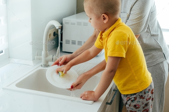 child boy helping mom with housework washing dishes in the kitchen sink. mommy teaches the child.