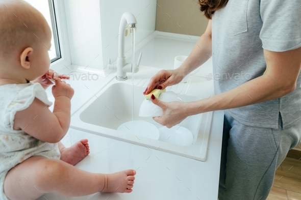 A woman washes the dishes in the kitchen sink. A baby is sitting on the table next to her.