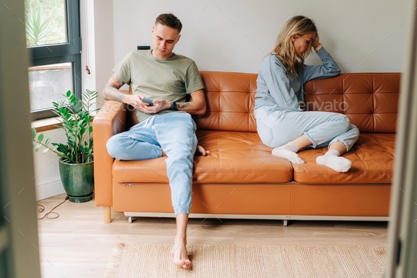 A quarreled couple is sitting on the couch, the man is using a mobile phone, the woman is offended.