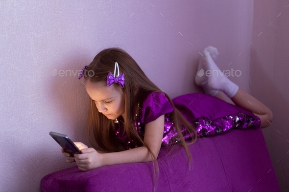 The girl is lying on the back of a purple armchair in a room with purple curtains and playing on the
