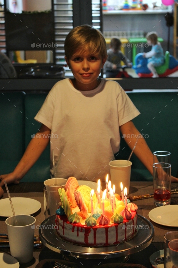 A boy makes a wish in front of a multicolored decorated cake with lit candles on his birthday