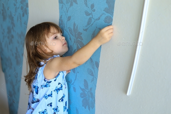 A cute little girl helps to make repairs in the apartment, tearing off old wallpaper from the wall