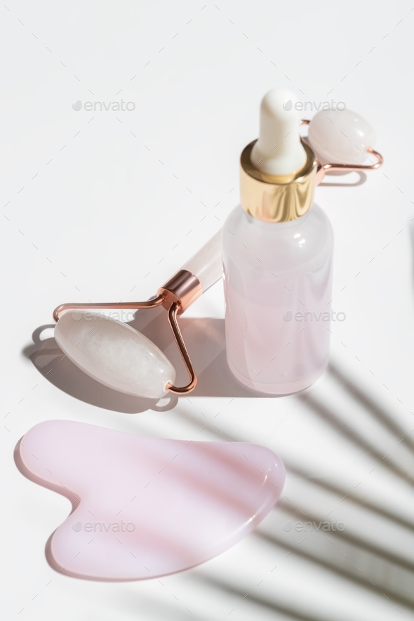 Pink Gua Sha facial massage tools. Rose Quartz jade roller and beauty facial oil on white background