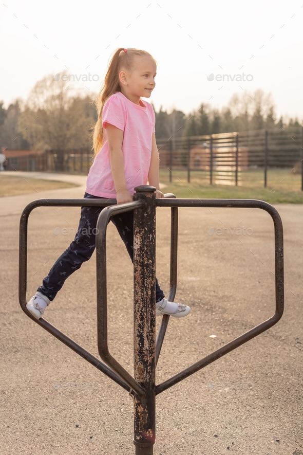 A small child girl with loose hair is spinning on a street horizontal bar at the stadium.
