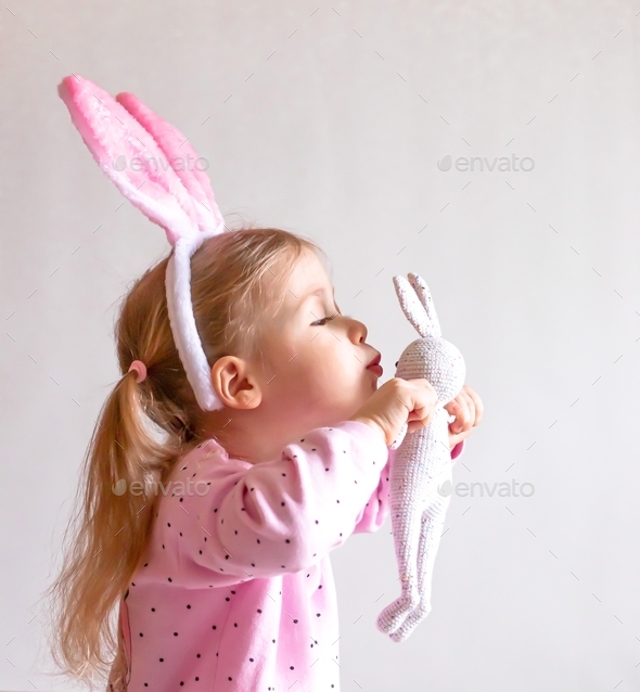Cute happy girl in a pink jacket with the Easter bunny ears kissing her toy rabbit
