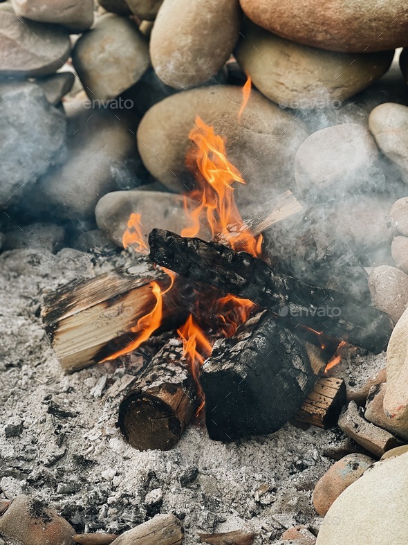 Burning wood in a smoky campfire made of rocks to protect it from the wind  - Stock Photo - Images