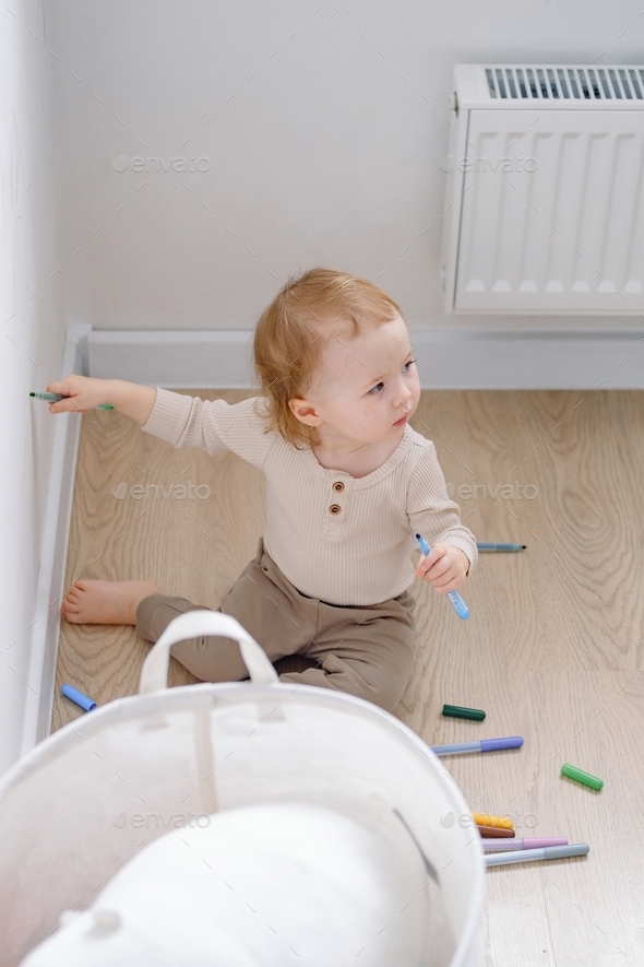 A toddler sitting by the white wall holding colored markers and drawing on the wall. Baby developing