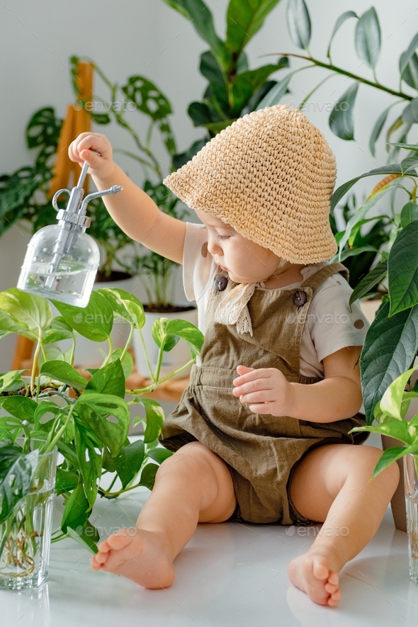 Baby and home jungle. A toddler sitting surrounded by potted room plants and holding water spray.