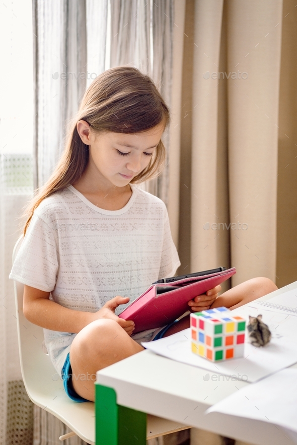 Caucasian school age girl sitting at the messed desk using tablet. Generation Z girl playing game.