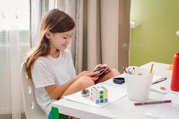 Caucasian school age girl sitting at the messed desk using tablet. Generation Z girl playing game.