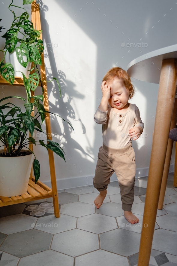 A cute toddler walking squinting from the sun with houseplants on the background. Learning to walk.
