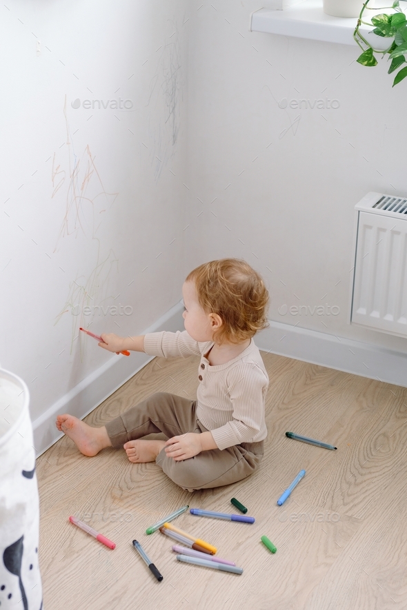 A toddler drawing on the white wall by colored markers. Allowing children to use walls to draw.
