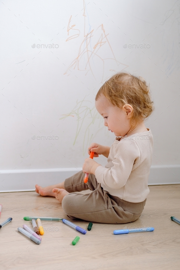A toddler holding colored marker and drawing on the white wall. Developing imagination and art.