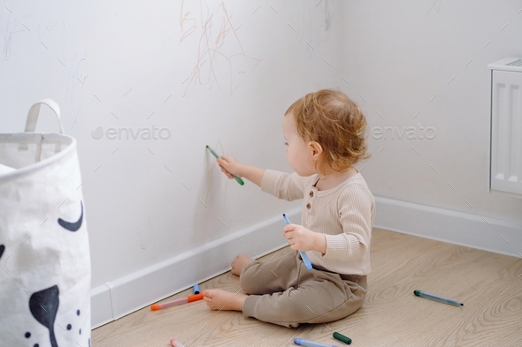A toddler drawing on the white wall using colored markers. Developing imagination.