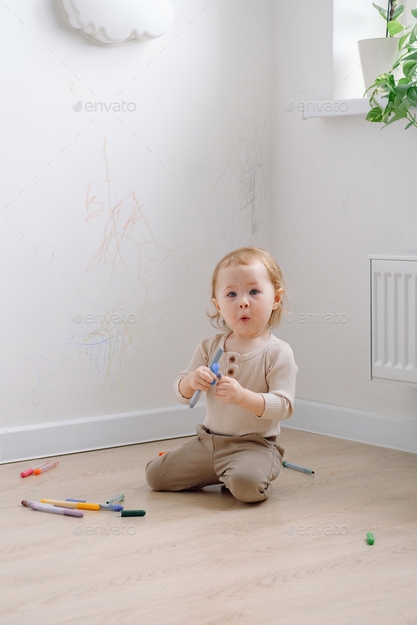A toddler making surprised face sitting by the white wall drawing on it by colored markers.