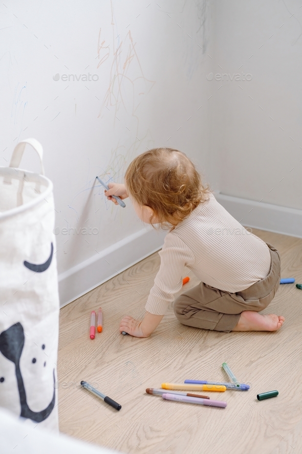 A toddler sitting on knees on the floor and drawing on the white wall by colored markers.