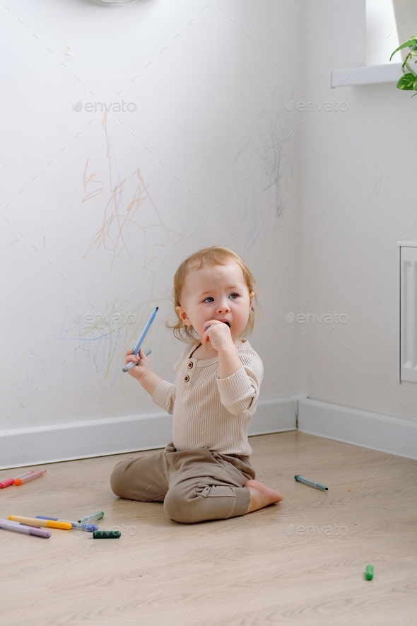 A toddler sitting by the white wall that has been drawn with colored markers and chewing a marker.