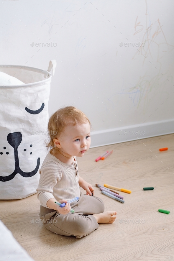 A toddler sitting by the white wall that has been drawn with colored markers and looking curiously.
