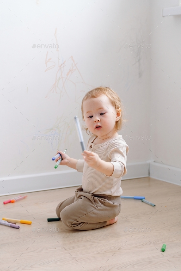 A toddler sitting by the white wall that has been drawn with colored markers and looking interested.