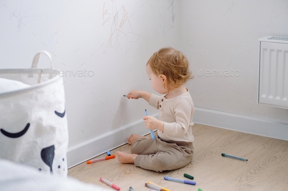 A toddler sitting by the white wall drawing on it with colored markers.