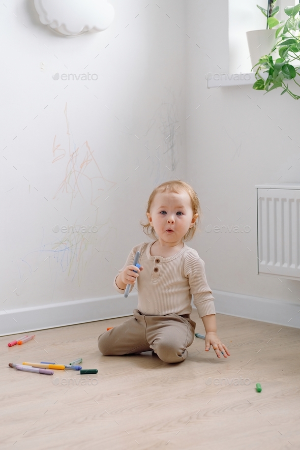 A toddler making surprised face sitting by the white wall drawing on it by colored markers.