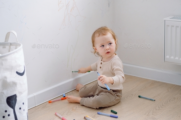 A toddler sitting by the white wall holding colored markers and drawing on the wall, looking away.