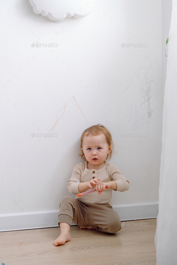 A toddler sitting by the white wall that has been drawn with colored markers and looking guilty.