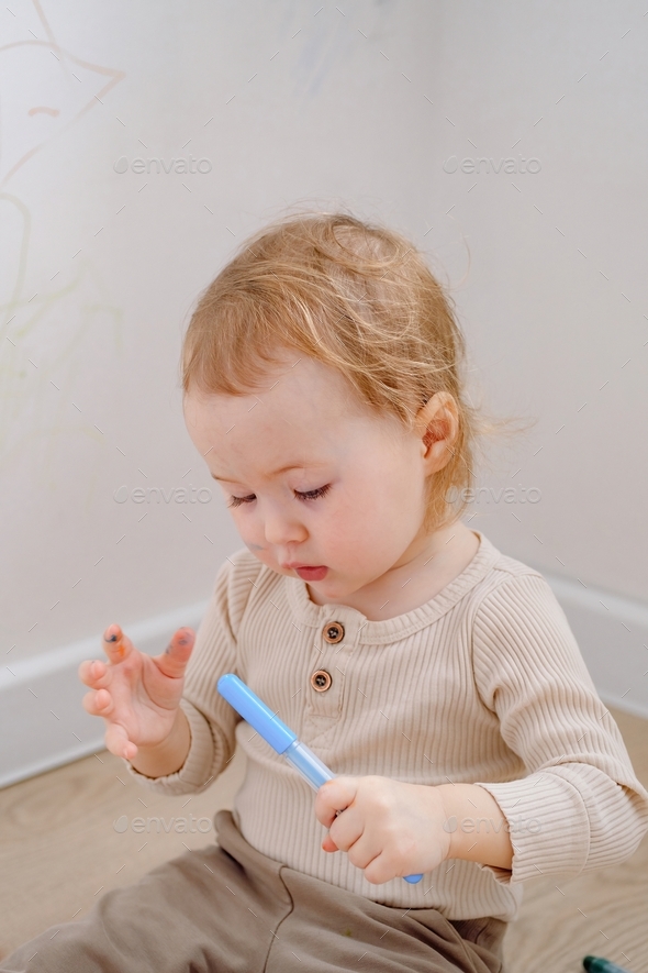 Close up of a cute toddler holding colored marker ready to draw on the white wall and himself.