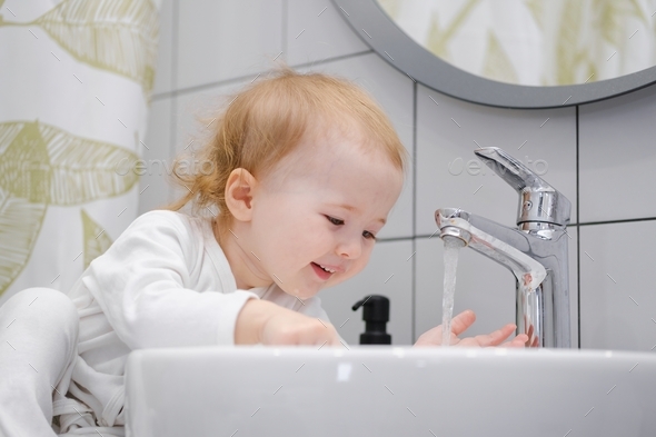 A cute smiling toddler washing hands under water in sink in the ...